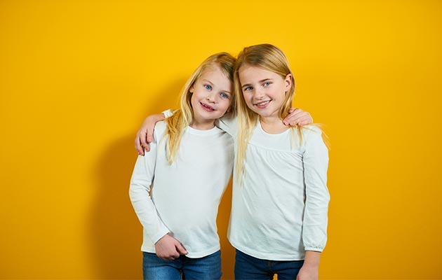 Zwei blonde Mädchen stehen Arm in Arm vor einem gelben Hintergrund