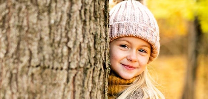 Mütze juckt? in kleines Mädchen mit einer Mütze auf dem Kopf, das hinter einem Baum um die Ecke schaut.
