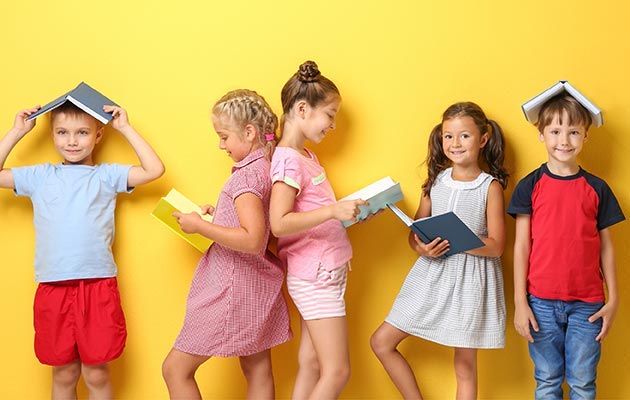 Eine Gruppe von Kindern steht vor einem gelben Hintergrund mit Büchern in der Hand und auf dem Kopf