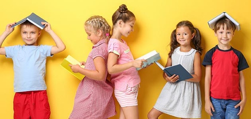 Eine Gruppe von Kindern steht vor einem gelben Hintergrund mit Büchern in der Hand und auf dem Kopf