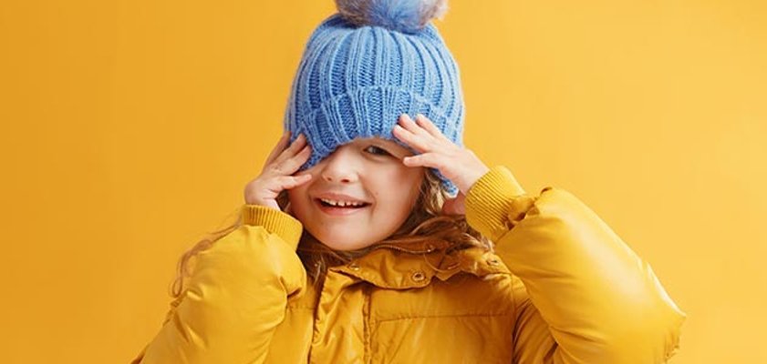 Winterzeit = Läusezeit? Ein kleines Kind vor einem gelben Hintergrund, mit einer gelben Jacke und einer blauen Mütze, richtet mit ihren Händen die Mütze.