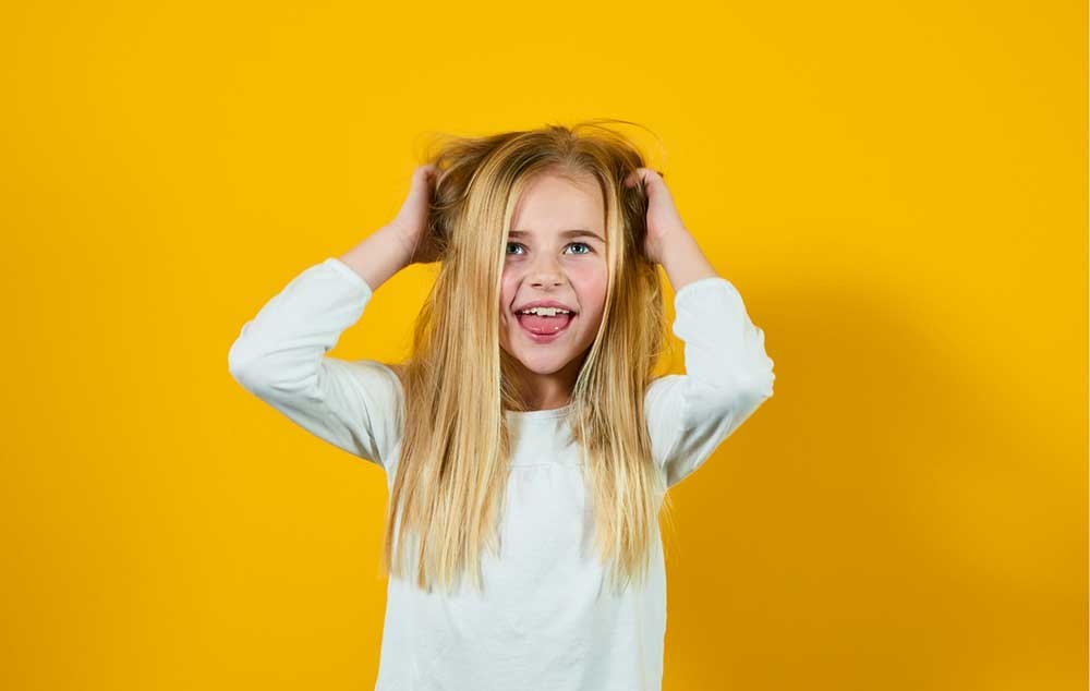 Schutz vor Kopfläusen Ein Mädchen steht vor einem gelben Hintergrund und packt sich in die Haare.